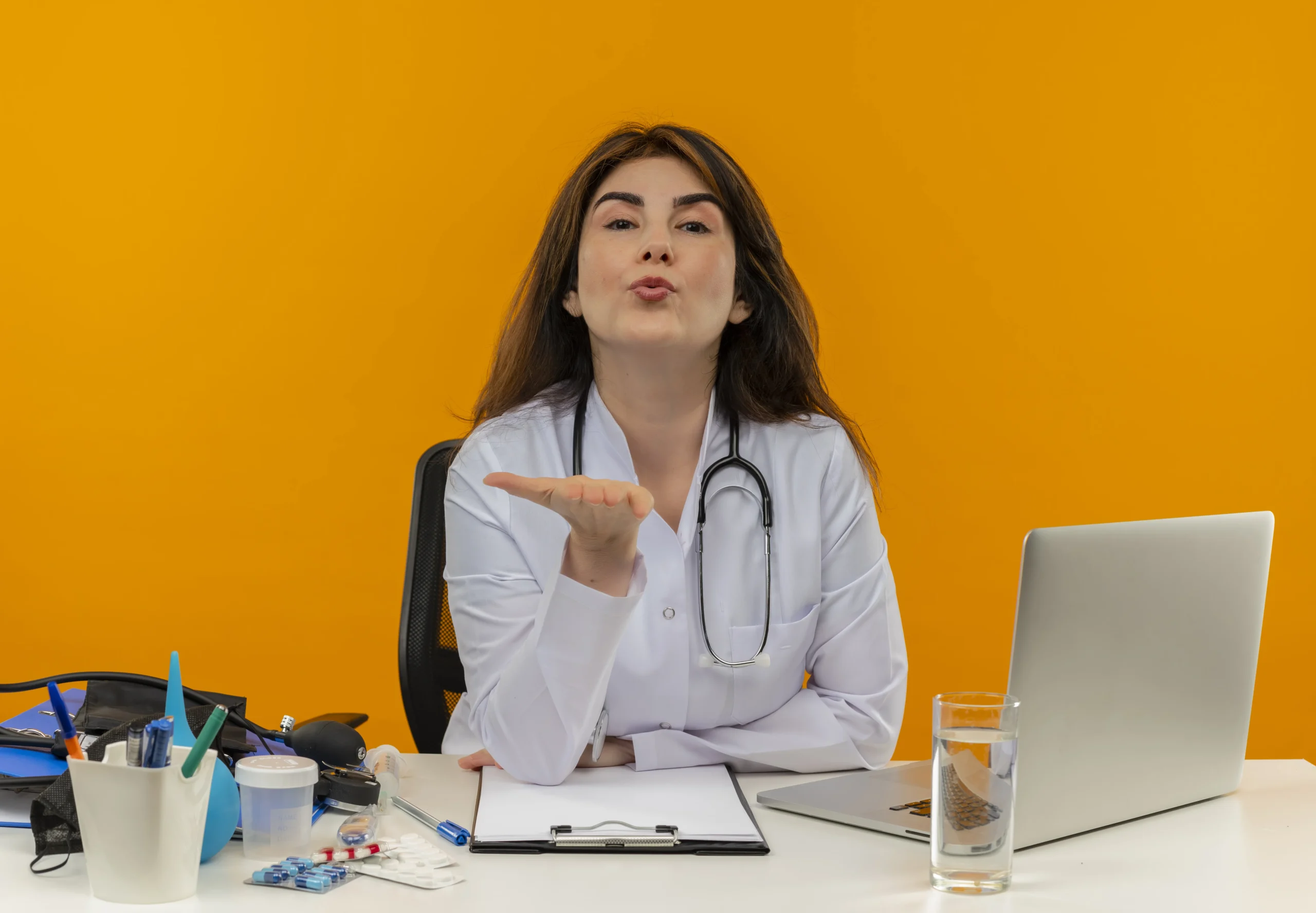 Female doctor sitting at her desk, sending a blow kiss, symbolizing care and support for women’s health, including AUB in women's health.