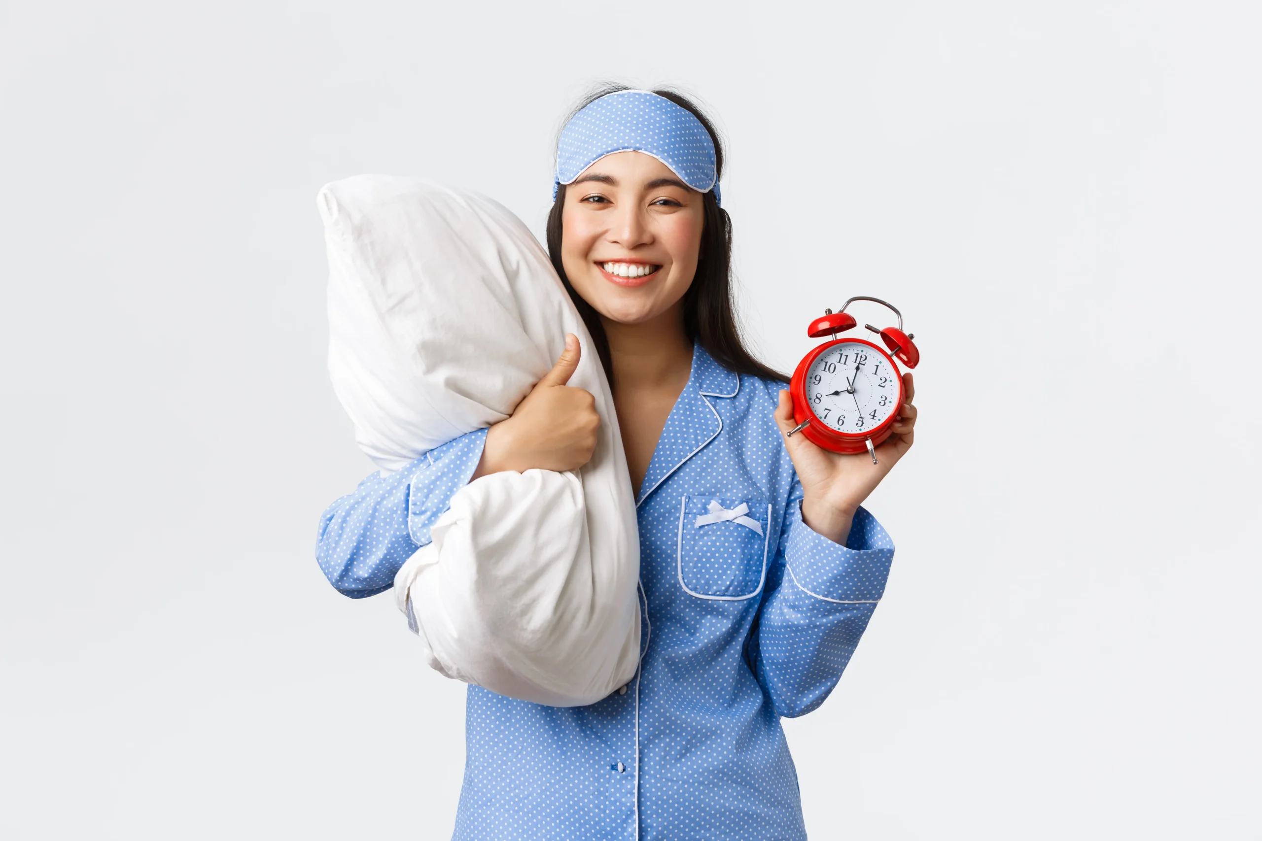 Happy woman in blue pajamas with a sleep mask, holding a pillow and alarm clock, highlighting how daily habits affect long term health, especially sleep habits.