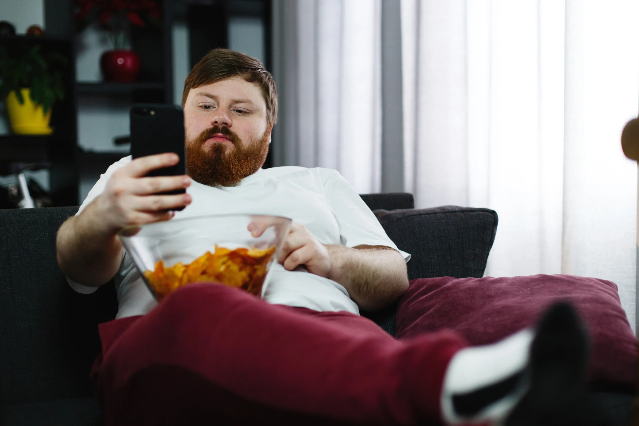 What is a Sedentary Lifestyle? A man sitting on a sofa using his smartphone while eating snacks, representing prolonged sitting, screen time, and physical inactivity.