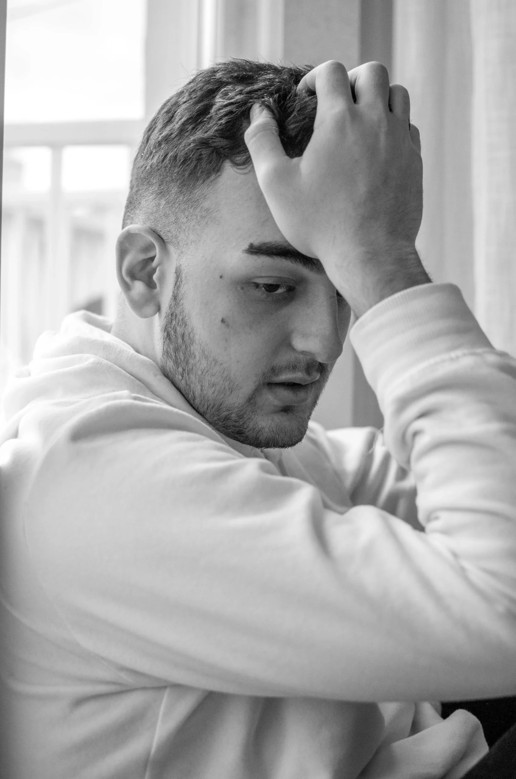 Young man sitting by the window, looking stressed and worried, symbolizing the impact of mental health stigma in men and its effect on emotional well-being.