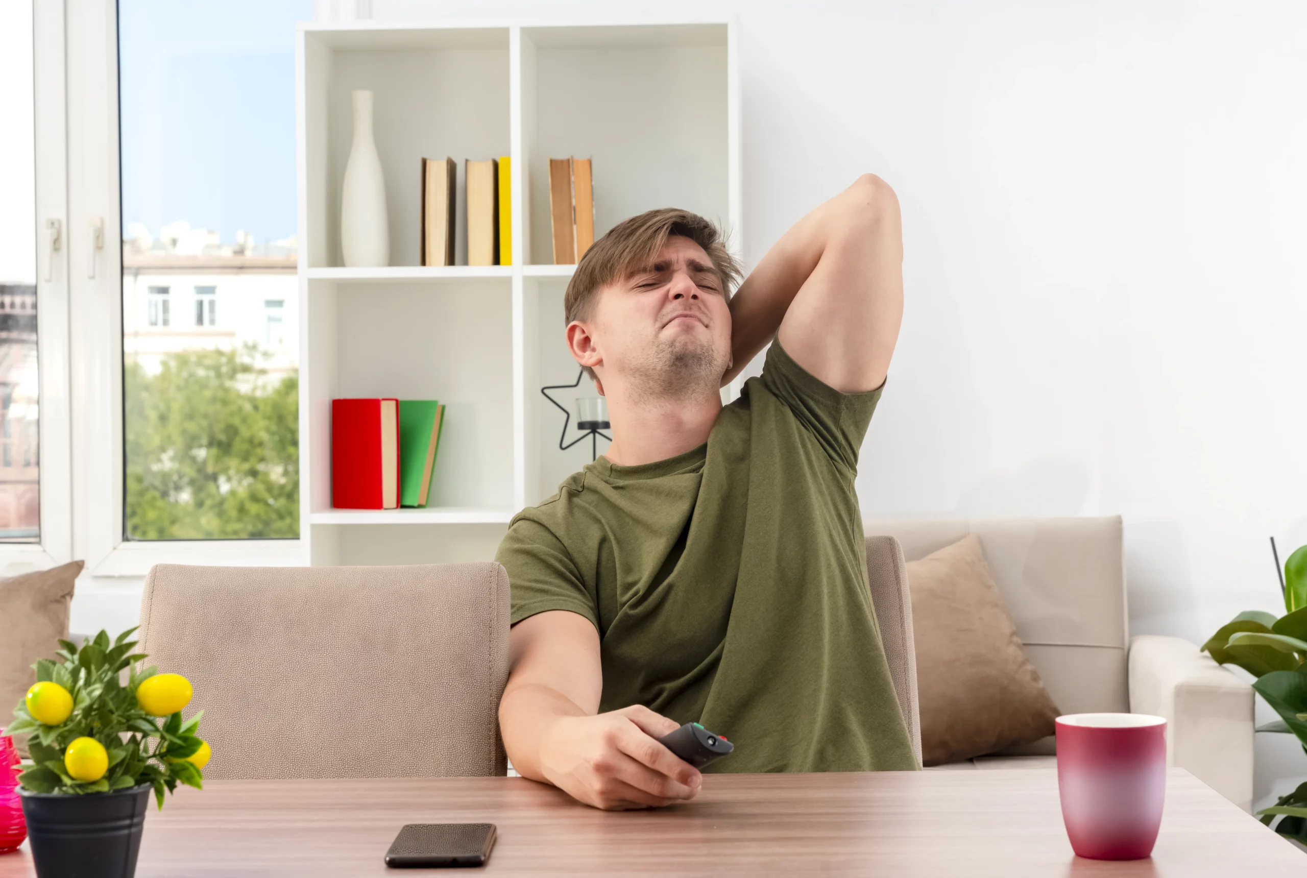 A man experiencing neck pain while sitting at a table, showing the early signs your body needs rest, as he holds a remote and appears fatigued in his living room.