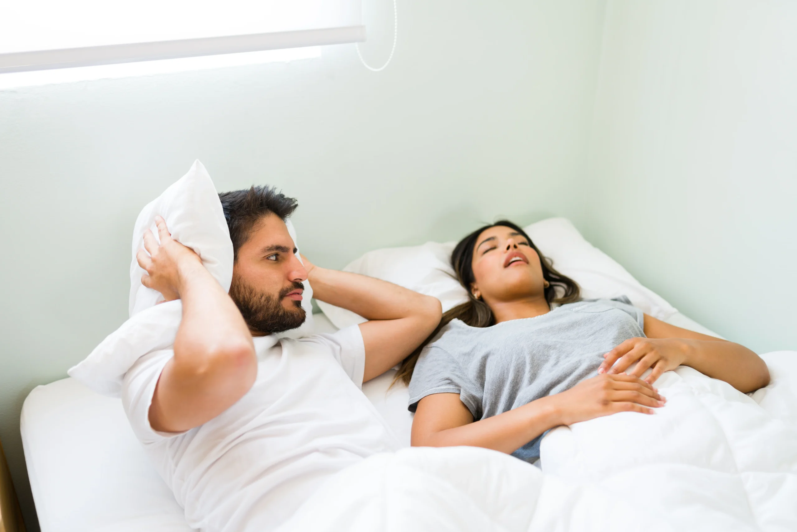 Man covering his ears with a pillow, frustrated by his girlfriend’s snoring, highlighting the impact of does snoring affect sleep quality? on relationships and sleep.