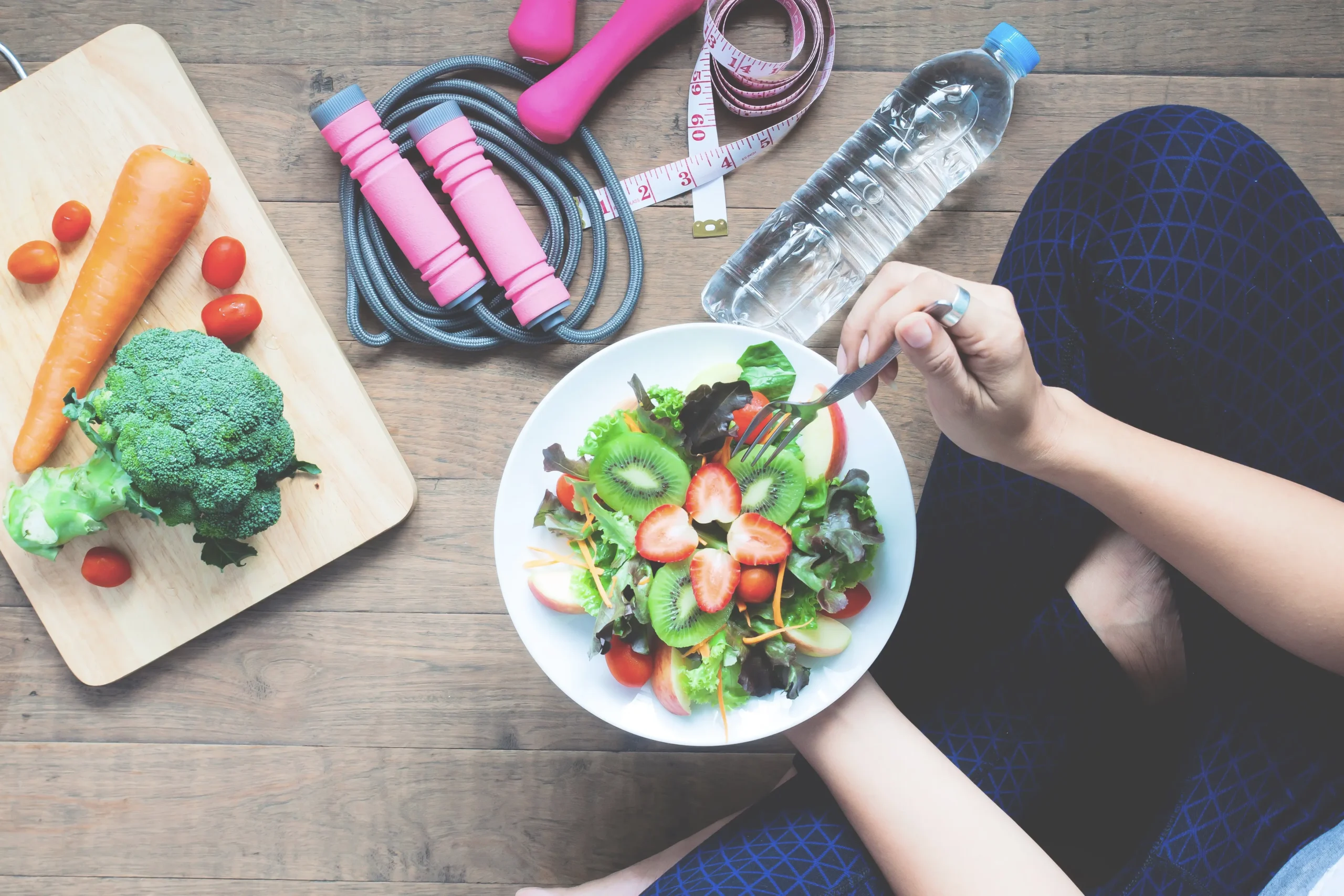 Woman eating a healthy salad with fresh vegetables like broccoli, tomatoes, and carrots, with fitness equipment like dumbbells, measuring tape, and water bottle, promoting healthy lifestyle habits.