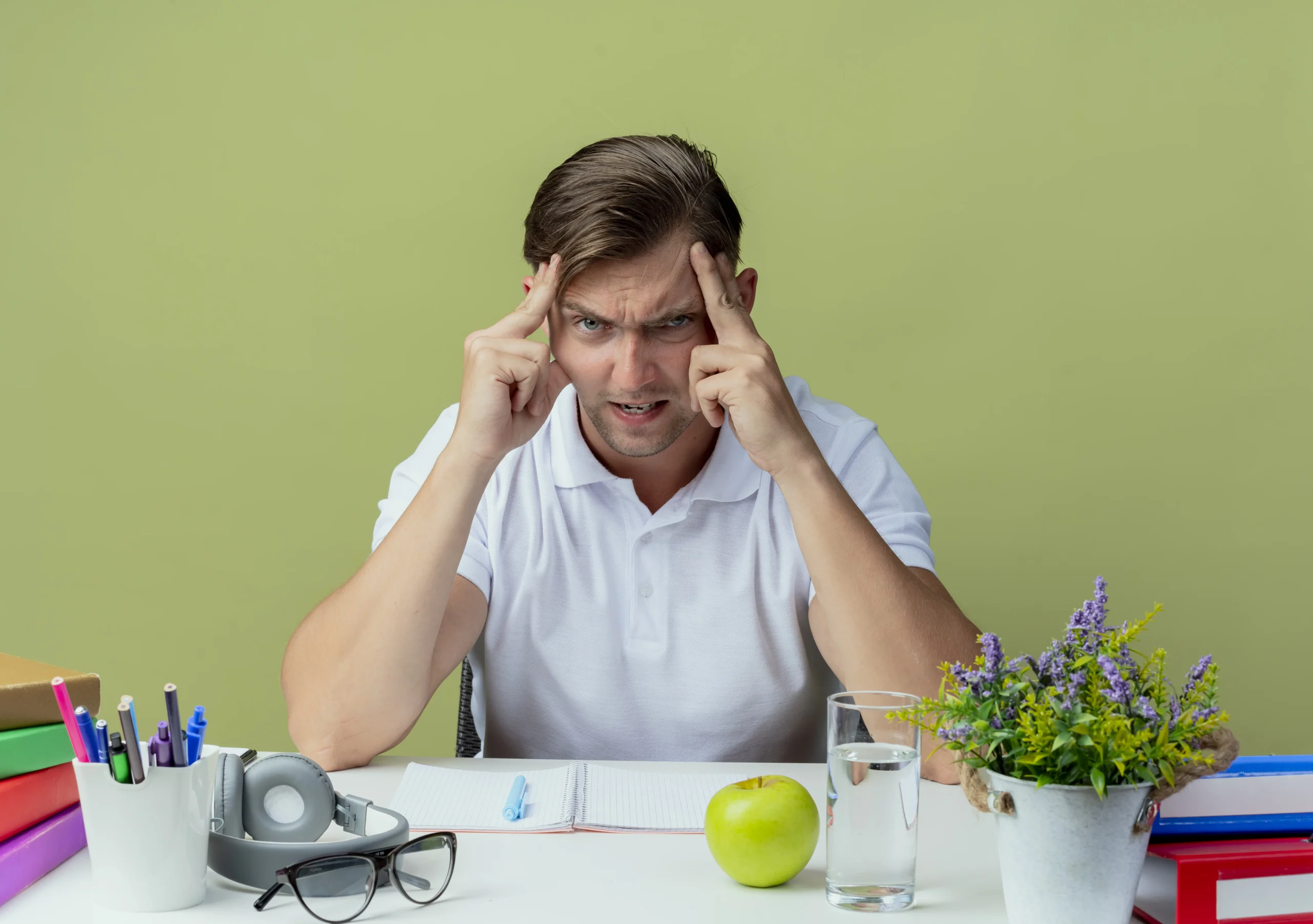 Frustrated man sitting at a desk with school supplies, holding his forehead, representing the effects of stress and how stress impacts physical health.